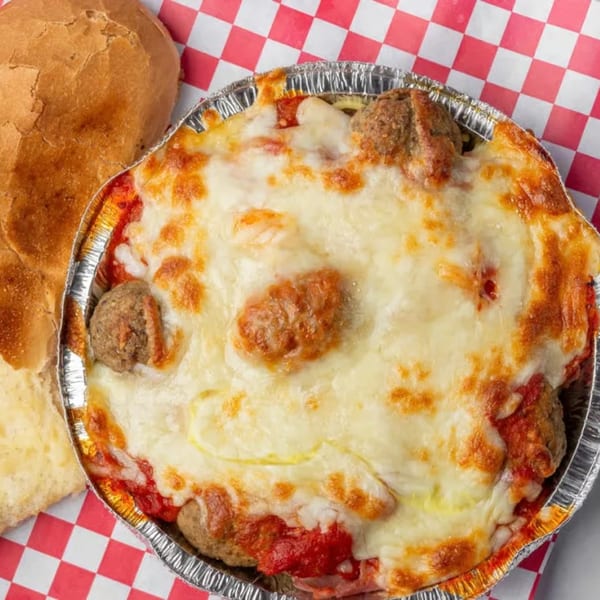 Baked pasta with meatballs and melted cheese in a foil pan, next to a bread roll.