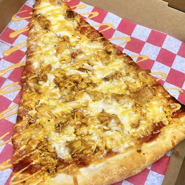 A large slice of Pascali's Pizza Corner pizza with melted cheese, chicken, and a creamy sauce drizzle.